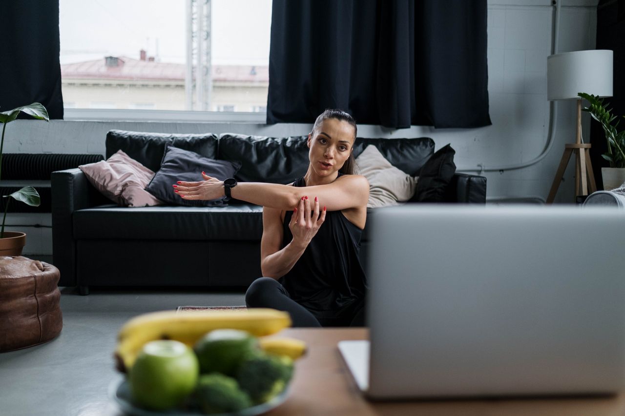 Femme faisant du yoga chez elle avec un ordinateur portable, étirant ses bras dans une ambiance chaleureuse de salon.